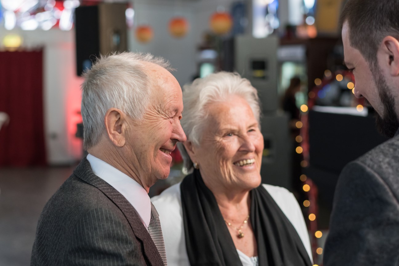 Ein Senior und eine Seniorin (gut angezogen) lachen einen Mann an